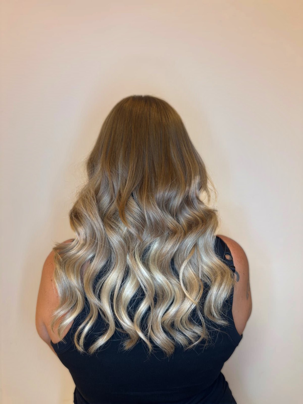 Woman with long honey-blonde AVRÉE hair extensions from behind, against an ivory wall